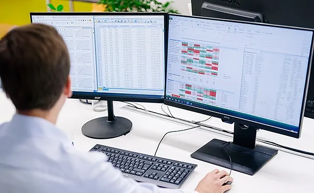 Thomas Schuster arbeitet an zwei grossen Computerbildschirmen, auf denen umfangreiche Excel-Tabellen mit Diagrammen und farblich markierten Daten zu sehen sind.