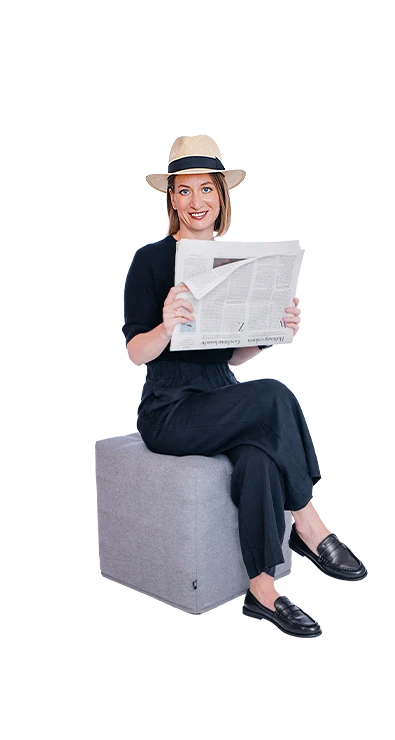 Mag. Barbara Khayat-Platzl, BSc (WU) sitzt mit übergeschlagenen Beinen und Zeitung in der Hand lächelnd auf einem Hocker – entspannt, stilvoll und immer gut informiert.