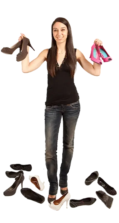 Mag. Raquel Herrero Pastor steht umgeben von einer Sammlung verschiedenster Schuhe und hält je ein Paar in jeder Hand - stilbewusst, entschlossen und voller Schuhleidenschaft.