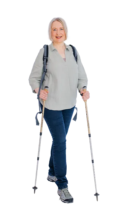Mag. Sonja Ehrenberger geht mit Wanderstöcken und Rucksack lächelnd auf die Kamera zu – naturverbunden, aktiv und voller positiver Energie.