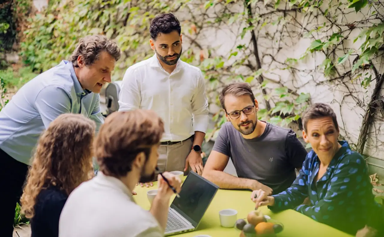 Gereon Friederes, Philipp Zelenka, Daniel Winzer, Dagmar Jiresch, and Youssef Kawmi are sitting at a table in the garden of the marketmind office together with clients, engaged in lively discussion. On the table are a laptop, coffee cups, and a bowl of fruit.