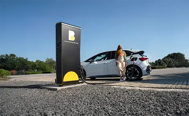A customer leans against an electric car connected to a modern charging station of Burgenland Energie and relaxes in the sun while waiting for the charging process.
