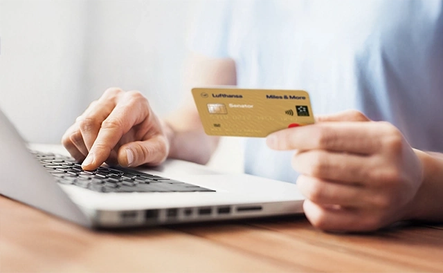 A person holds a golden Lufthansa Miles & More credit card in hand and types on a laptop with the other hand.