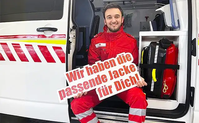 A volunteer paramedic in red emergency clothing sits in the open side door of an ambulance holding a sign reading 'We have the right jacket for you!' In the background, equipment is visible in the vehicle.