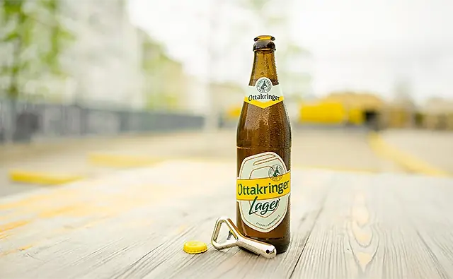 Opened bottle of Ottakringer Lager beer with crown cap and bottle opener on a wooden table outdoors.