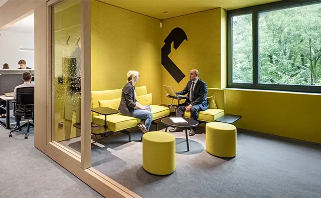 Consultation between bank advisor and customer in a modern, yellow-designed meeting room with large windows and view into greenery. In the background, an open office space with several working people is visible.