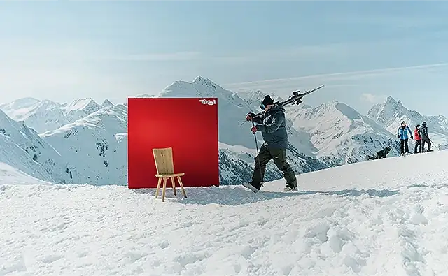 Scene from the target group-specific advertising campaign 'Always there for you'. Skier with skis on his shoulder walking past a 'Tirol' advertising board on the ski slope, in front of which stands a traditional wooden chair. In the background, other skiers resting and a beautiful mountain panorama.