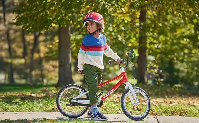 Child with red bicycle helmet stands with a red Woom children's bicycle in the park.