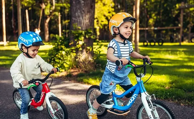 Two children riding colorful Woom balance bikes happily through a sunny park. Symbolic image for child-appropriate mobility, safe riding pleasure, and thoughtful product design by the Woom brand.