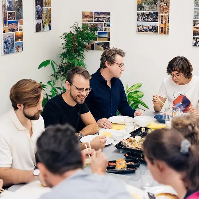 Several colleagues are sitting together at a table eating sushi while engaged in lively conversation. Philipp Zelenka, Daniel Winzer, Gereon Friederes, and Karin Graf can be seen in the foreground. Green plants and vacation photos on the wall can be seen in the background.