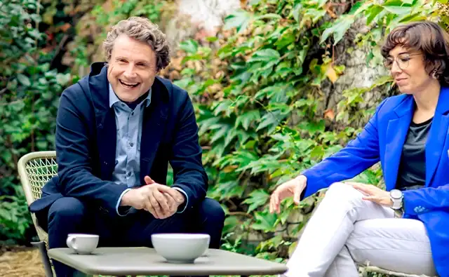 Gereon Friederes sits smiling in a garden chair and engages in conversation, while Karin Graf, wearing a blue blazer, sits opposite him and communicates intently with gestures. Between them, coffee cups stand on a table.