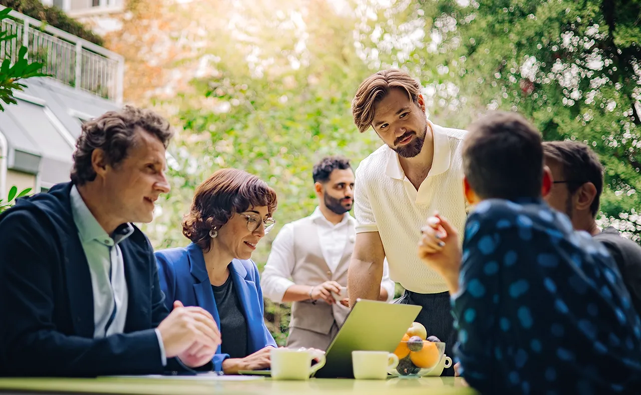 Gereon Friederes, Karin Graf, and Philipp Zelenka are sitting at a table in the garden of the marketmind office together with clients, engaged in lively conversation. On the table are a laptop, coffee cups, and a bowl of fruit. Youssef Kawmi can be seen in the background.