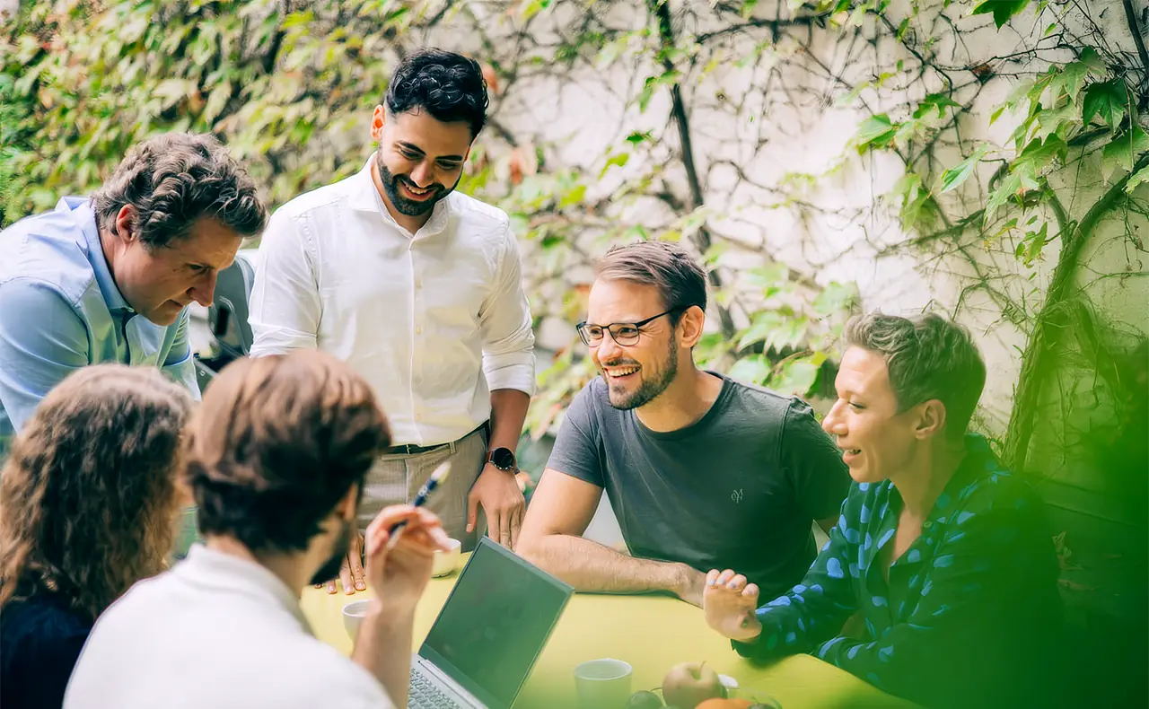 Gereon Friederes, Philipp Zelenka, Daniel Winzer, Dagmar Jiresch, and Youssef Kawmi are sitting at a table in the garden of the marketmind office together with clients, engaged in lively discussion. On the table are a laptop, coffee cups, and a bowl of fruit.