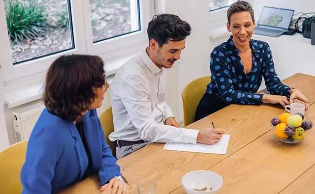 Karin Graf, Dagmar Jiresch, and Jakob Schinagl are sitting at a wooden table in a bright meeting room. They are chatting cheerfully. Jakob Schinagl is taking notes on a piece of paper. There is fruit and a coffee cup on the table.