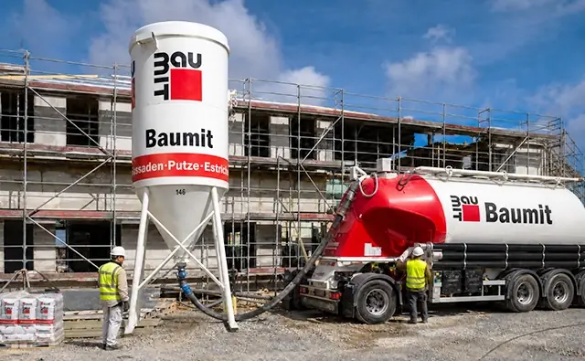 Baumit silo and Baumit truck on a construction site in front of a scaffolded building while workers process building material. Image represents construction materials, building processes, and professional façade and plaster solutions.