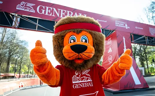 Generali mascot lion in red costume shows thumbs up at a running event.