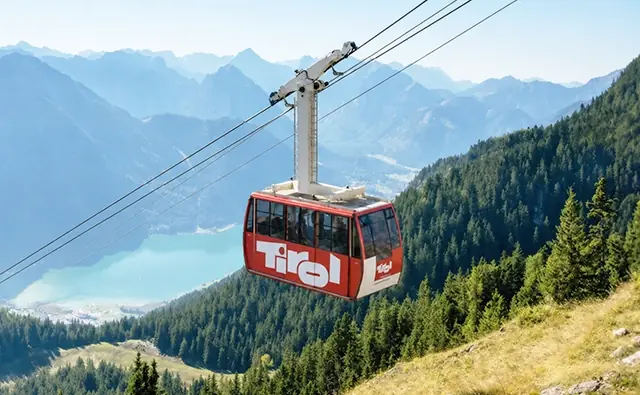 A cable car with Tirol branding during the mountain ascent - with a view of a turquoise mountain lake and forested Alps.