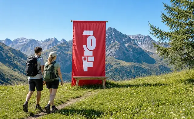 Two hikers stand on an alpine meadow in front of a red Tirol banner with a mountain landscape in the background. Image represents nature, tourism, and mountain experiences in Tirol.