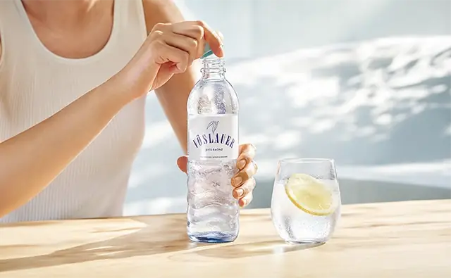 Person opening a bottle of Vöslauer sparkling mineral water next to a glass of water with a lemon slice.