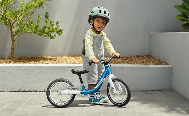 Child wearing a helmet stands with a blue woom balance bike in a sunny courtyard. Image represents children’s bikes, safe mobility, and playful learning to ride.