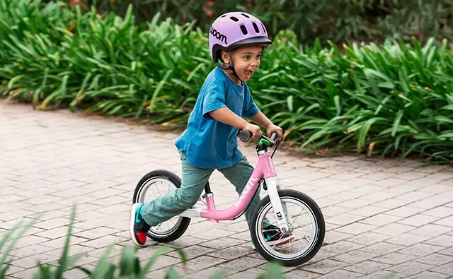 Child wearing a helmet rides a pink woom balance bike along a paved path in a green outdoor setting. Image represents children's bikes, safe mobility, and targeted brand communication.
