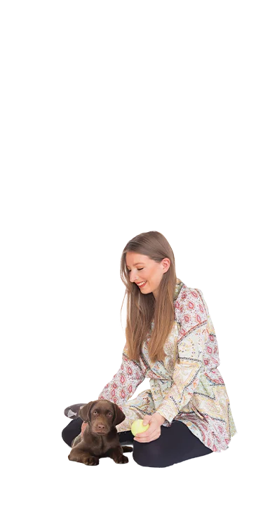 Caroline Peinitz-Kichler kneels on the floor with a small Labrador puppy, a yellow tennis ball in hand - animal-loving and warm-hearted.