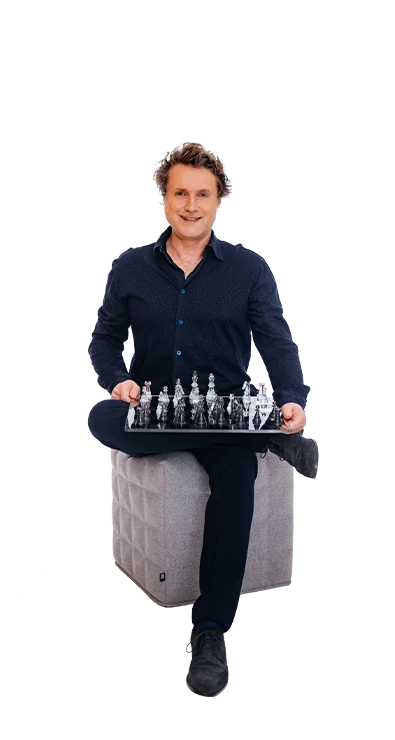 Dr. Gereon Friederes sits relaxed on a gray stool and holds a chessboard with transparent pieces - strategic, focused, and with a smile.