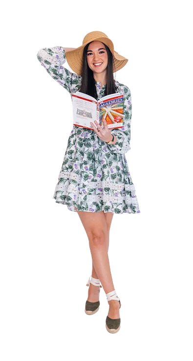 Giselle Kaufmann wears a floral summer dress and sun hat while smiling with a Madrid travel guide, awakening wanderlust.