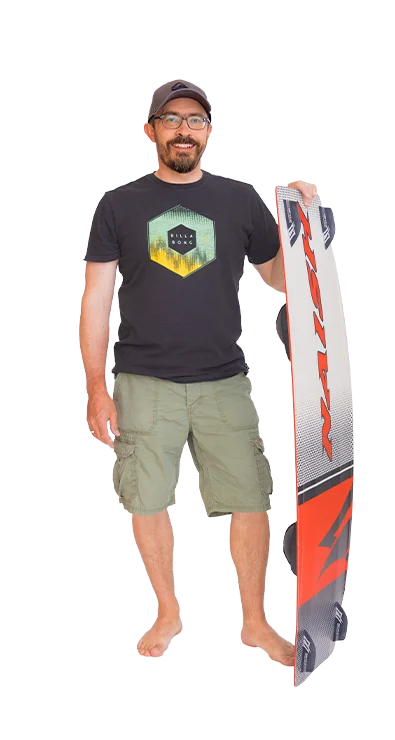 Gerhard Schüller stands barefoot, wears a T-shirt with a surf motif, and holds a kiteboard - athletic, relaxed, and ready for wind and waves.