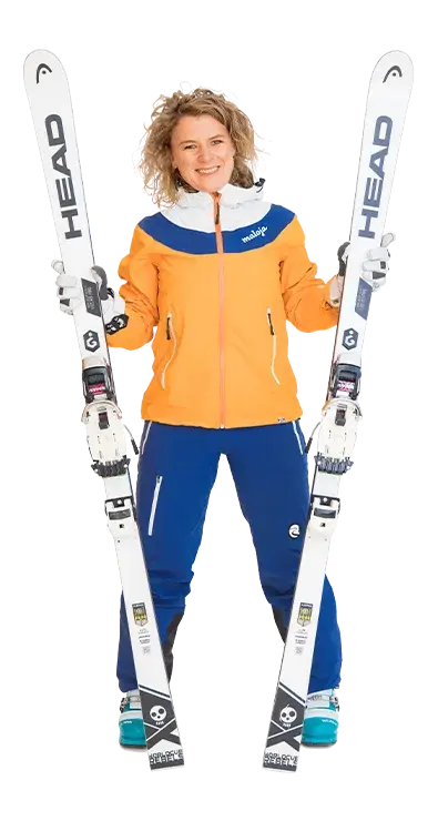 Heidi Thyri stands in complete ski gear with an orange-blue jacket and firmly holds two white skis in her hands - ready for the slopes and plenty of snow adventures.