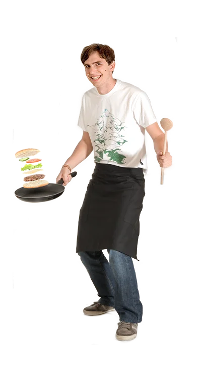 Manuel Nussbaumer wears a cooking apron, juggles a stacked burger in a pan and holds a cooking spoon in the other hand - creative, playful, and passionate about cooking.