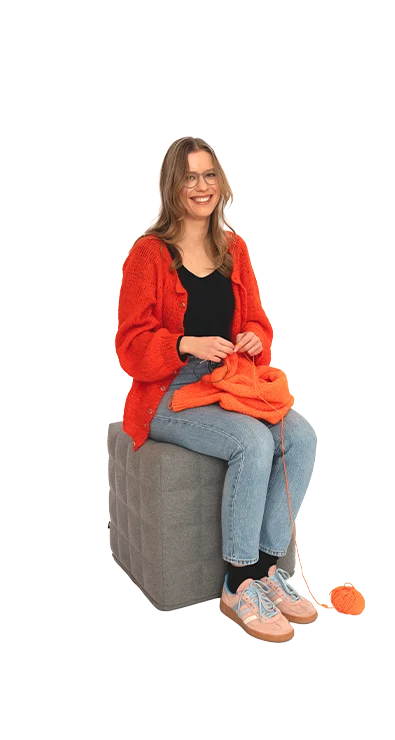 Polina Yaroshchuk sits on a stool with orange cardigan and wool, knitting materials in hand - creative, focused, and craftily skilled.