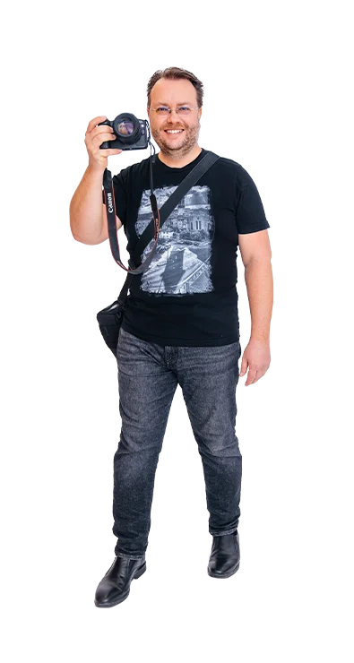 Dr. Wolfgang Rejzlik holds a camera ready to shoot and seems about to take a photo - equipped with shoulder bag and big smile.